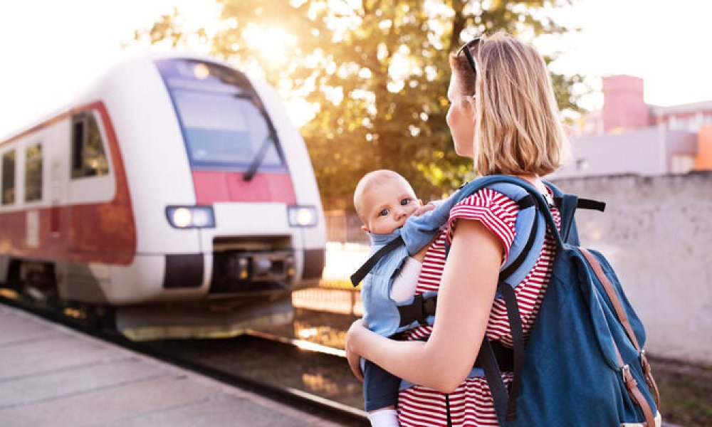 Mor med en baby i bæresele som står på en togstasjon og ser på toget som kommer.