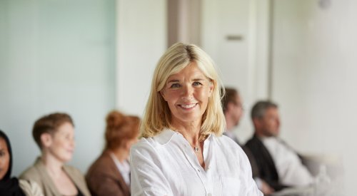 Illustrasjonsfoto: Dame i hvit skjorte og armene i kryss smiler til kamera, bak sitter flere mennesker. 