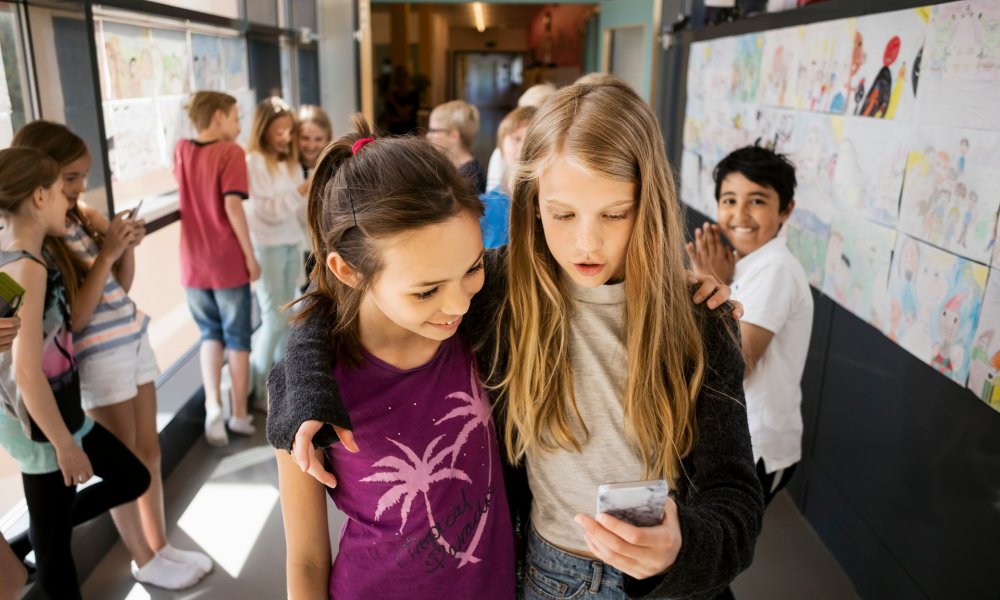 Illustrasjonsfoto: To barn holder rundt hverandre i gangen på en skole. Den ene jenta holder en mobil.