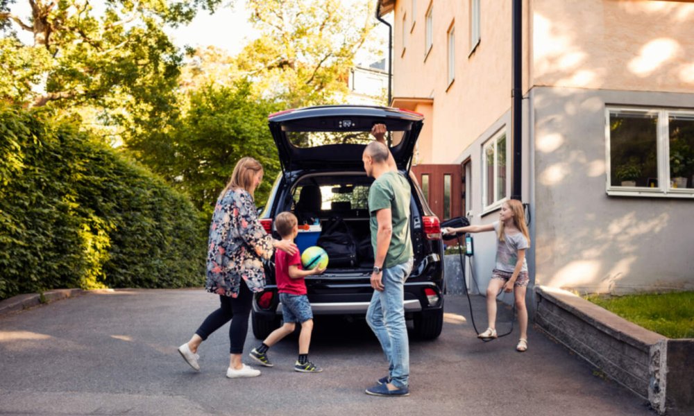 Familie med mor, far og to barn pakker bilen utendørs om sommeren. Jenta lader bilden, og gutten har en ball i hånden. 