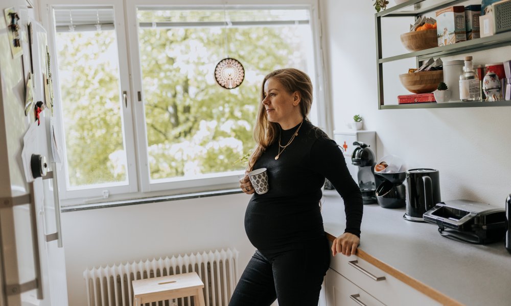 Illustrasjonsfoto: En gravid kvinne lener seg mot kjøkkenbenken med en kopp i hånden.