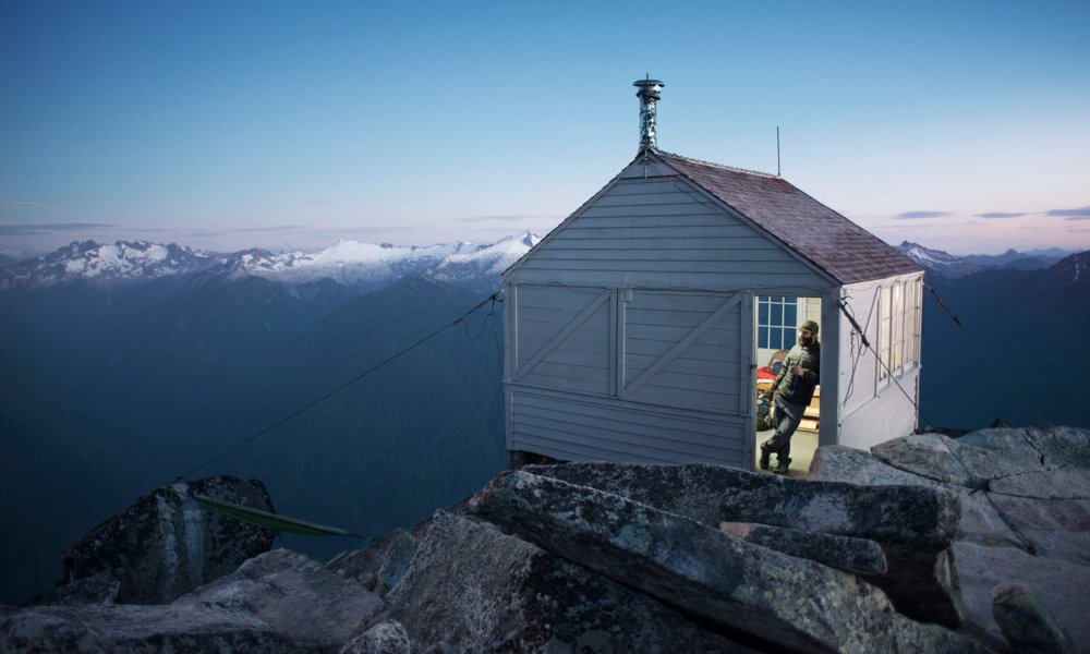 Illustrasjonsfoto: En mann står og lener seg i dørkarmen til en hvit hytte på fjellet.